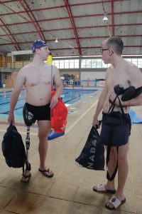 Lucas e Nelson conversam de pé, ao lado da piscina, com roupas de banho e chinelo. Lucas usa uma prótese de perna